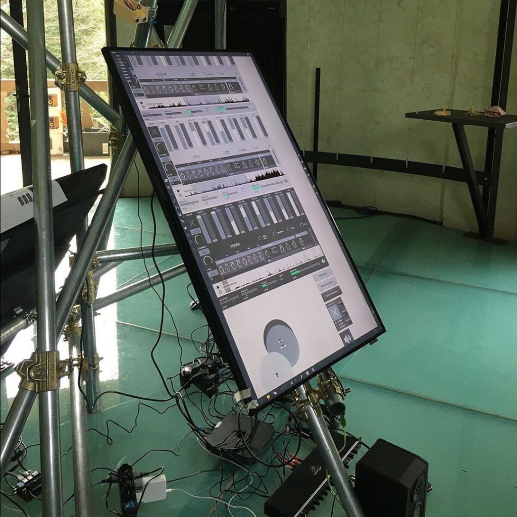 A large touchscreen monitor displaying a digital audio interface, surrounded by cables and electronic equipment in a studio-like environment.